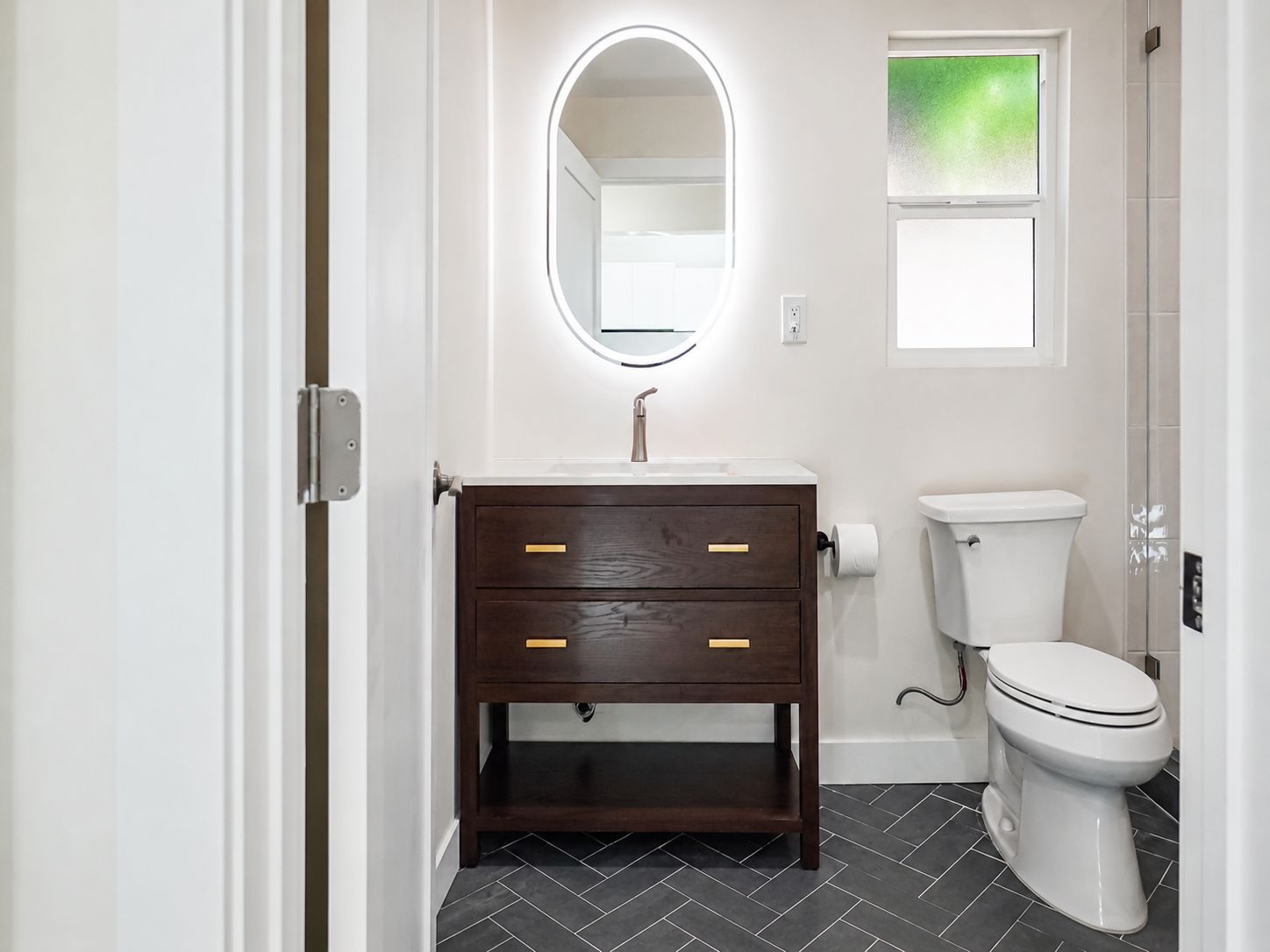 Bathroom vanity with backlit oval mirror and herringbone tile floor